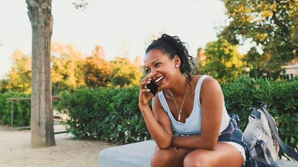 Laughing young mixed race woman sitting on park bench talking on cellphone