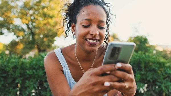 Laughing young mixed race woman sitting on park bench using cellphone