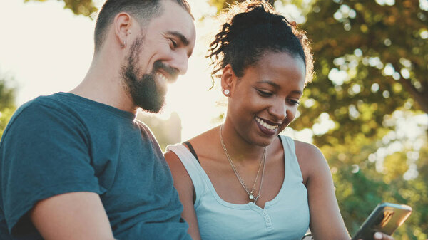 Happy interracial couple talking while sitting on bench, using cellphone