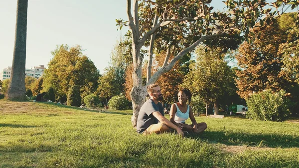Щаслива Усміхнена Міжрасова Пара Розмовляє Сидячи Траві Парку Підсвічування — стокове фото