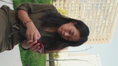 Genç kız modern binaların arka planında otururken akıllı telefon kullanıyor.