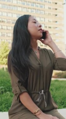 Modern binaların arka planında otururken akıllı telefondan konuşan genç bir kız.