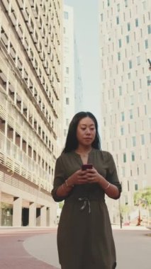 Modern binaların arka planında dururken cep telefonu kullanan genç, mutlu iş kadını.