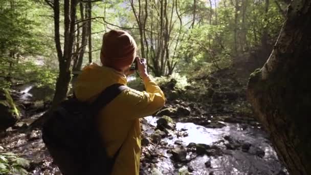 Fotografo barbuto in occhiali e impermeabile nella foresta di legno di montagna vicino al fiume. — Video Stock