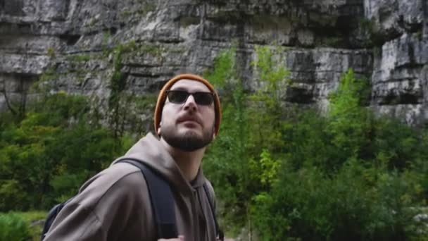 Viajante masculino com mochila em óculos de sol caminha para terras altas por estrada de montanha. — Vídeo de Stock