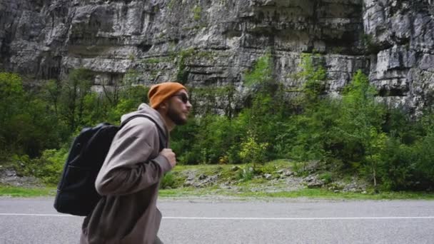 선글라스를 찬 가방을 든 남자 여행자 가산 길을 따라 고지대로 걸어가고 있다. — 비디오