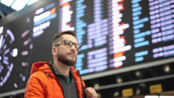 Lidé na letištním terminálu. Muži v brýlích s batohem procházky pod tabulí. — Stock video