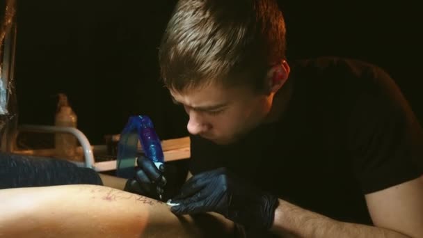 Tatueringskonstnär arbetar på kundben under lampa ljus i mörkt rum. Tatuerare butik — Stockvideo
