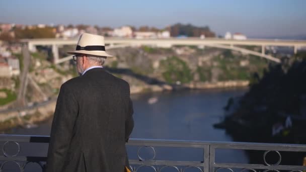 Vieux voyageur aux cheveux gris en chapeau de fedora blanc marche sur le Ponte de Dom Luis I. — Video
