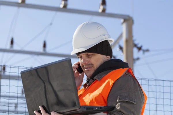 Electricista con PC y teléfono celular cerca de la subestación eléctrica —  Fotos de Stock