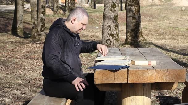 Man reading a book outdoors on a bench near table — Stock Video