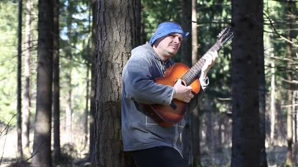 Mann spielt im Wald Gitarre und lehnt sich an Baum — Stockvideo