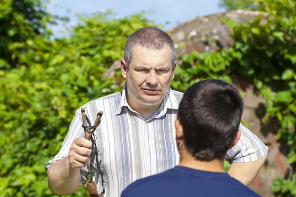 Angry man with slingshot in hand against the boy in summer — Stock Photo, Image