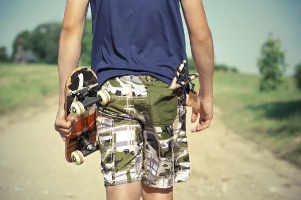 Jongen met skateboard en katapult in zak op landelijke weg in de zomer — Stockfoto