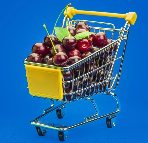 Carrinho de compras com cerejas frescas em um fundo azul — Fotografia de Stock