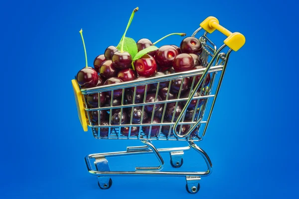 Carrito de compras con cerezas frescas sobre fondo azul — Foto de Stock