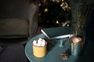 Koyu yeşil oda Noel 'de, ağaç sandalyeli marshmallow masasında sarı bardakta içecek. 