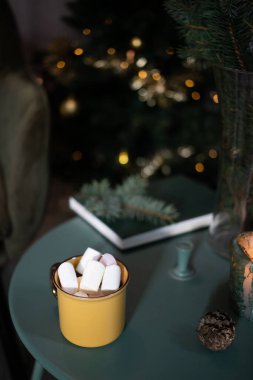 Koyu yeşil oda Noel 'de, ağaç sandalyeli marshmallow masasında sarı bardakta içecek. 