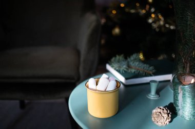 Koyu yeşil oda Noel 'de, ağaç sandalyeli marshmallow masasında sarı bardakta içecek. 