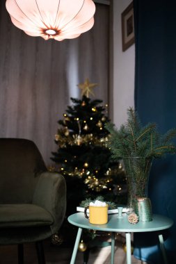 Koyu yeşil oda Noel 'de, ağaç sandalyeli marshmallow masasında sarı bardakta içecek. 