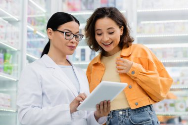 Eczanedeki neşeli müşterinin yanında dijital tablet kullanan gözlüklü Asyalı eczacı 