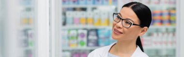 smiling asian pharmacist in glasses looking away in drugstore, banner clipart