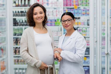 Eczanede gülümseyen hamile bir kadının elini tutan gözlüklü mutlu Asyalı eczacı. 