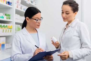 Beyaz önlüklü eczacı, Asyalı meslektaşının yanında gözlüklü, eczanede panoya yazı yazıyor. 