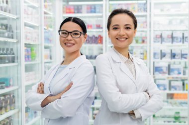 Beyaz önlüklü neşeli ırklar arası eczacılar eczanede çapraz kollarla dikiliyorlar.