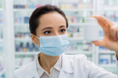 Tıbbi maskeli esmer eczacı eczanedeki bulanık şişeye bakıyor.