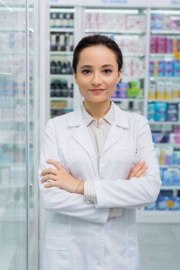brunette pharmacist looking at camera while standing with crossed arms in drugstore clipart