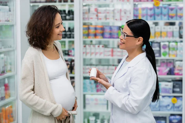 在药店里 亚洲药剂师戴着眼镜 手里拿着装有维生素的瓶子 旁边站着快乐的孕妇 — 图库照片