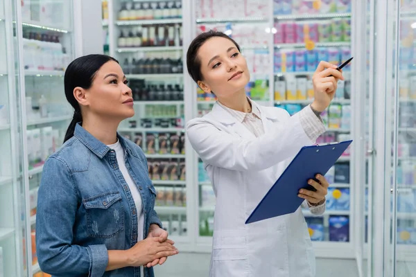 药剂师在药店拿着写字板时 用笔尖指向亚洲妇女 — 图库照片
