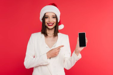 Noel Baba şapkalı neşeli kadın kırmızı ekranda boş ekranlı akıllı telefonu işaret ediyor. 