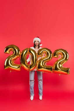 Noel Baba şapkalı genç kadın kırmızı arka planda 2022 numaralı balonların yanında duruyor.