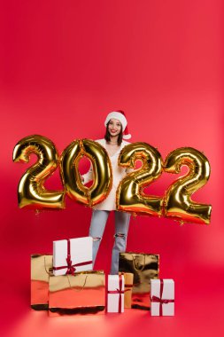 Noel Baba şapkalı mutlu kadın 2022 numaralı balonların yanında duruyor. Kırmızı arka planda hediyeler ve alışveriş torbaları. 