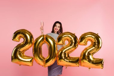 Gülen kadın 2022 model balonların yanında elinde şampanya kadehi tutuyor. Pembe renkte izole edilmiş. 