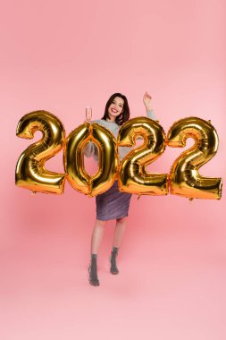 Güzel ve şık bir kadın, 2022 model balonların yanında pembe arka planda şampanya tutuyor.