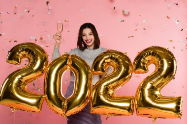 2022 model balonların yanında şampanya tutan neşeli bir kadın ve pembe arka planda konfeti.