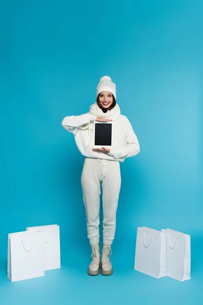 穿着温暖针织衣服的快乐女人 在蓝色背景的购物袋附近拿着数码平板电脑 — 图库照片