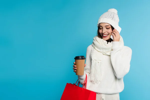 穿着保暖衣服的女人微笑着 一边用智能手机交谈 一边拿着购物袋和咖啡 用蓝色把自己隔离起来 — 图库照片