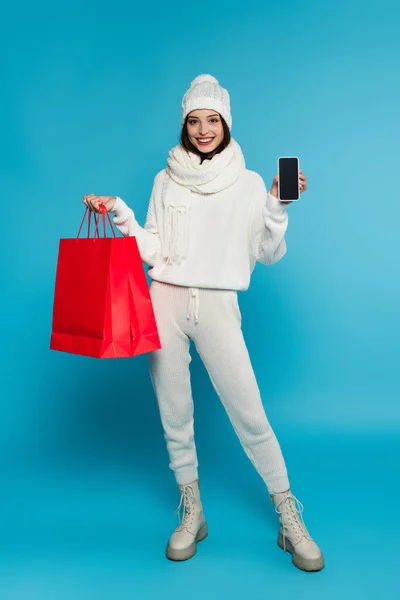 穿着针织服装的女人笑着 展示智能手机上的空白屏幕和蓝色背景的购物袋 — 图库照片