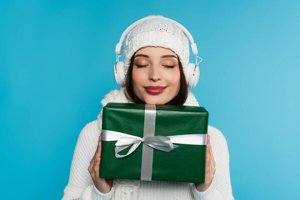 Woman with closed eyes in headphones and warm clothes holding gift box isolated on blue 