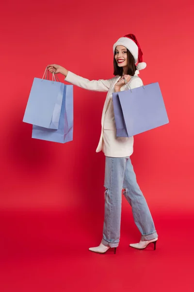 Veselá Žena Bundě Santa Klobouk Drží Nákupní Tašky Červeném Pozadí — Stock fotografie