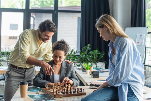 Smiling businessman pointing with finger at chess near interracial colleagues in office 