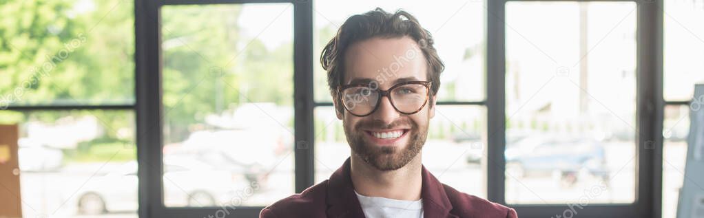 Cheerful businessman in eyeglasses looking at camera in blurred office, banner 