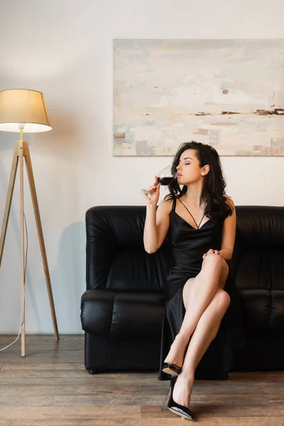 seductive woman in black slip dress holding glass and drinking wine while sitting on sofa