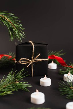 wrapped gift box near fir branches and candles with blurred red ribbon on black background clipart