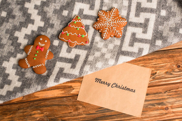 top view of gingerbread cookies on grey blanket with ornament near greeting card with merry christmas lettering on wooden surface 