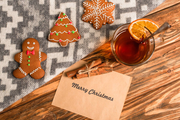 top view of gingerbread cookies on blanket near greeting card with merry christmas lettering and cup of tea on wooden surface 